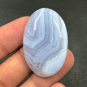 Può includere: Pietra preziosa ovale in agata blu pizzo con motivi a vortice blu e bianchi. La pietra lucida è tenuta in mano. Le dimensioni della gemma non sono visibili, ma probabilmente in centimetri.