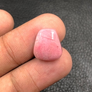 Puede incluir: Una gema rosa pulida con una forma redondeada e irregular. La piedra tiene una superficie lisa con sutiles variaciones de color y veteado. La gema se sostiene entre dos dedos, mostrando su tamaño y textura.