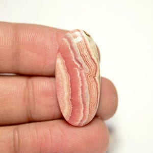 Puede incluir: Una piedra preciosa ovalada en cabujón con bandas rosadas y blancas. La piedra tiene una superficie lisa y pulida y se sostiene en una mano.