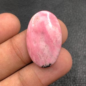 Puede incluir: Gema ovalada rosa con superficie pulida. La piedra tiene una mezcla de tonos rosa claro y oscuro con algunas inclusiones más oscuras. La gema se sostiene en una mano, mostrando su tamaño y forma. La piedra mide aproximadamente 3,8 cm de largo.