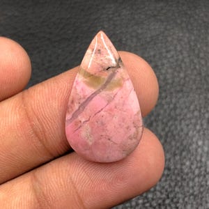 Puede incluir: Una gema pulida en forma de lágrima. La piedra presenta una mezcla de tonos rosas, marrones y beiges, con un color base rosa dominante. Se ven vetas y patrones en toda la piedra, lo que realza su aspecto natural. La gema se sostiene en una mano.