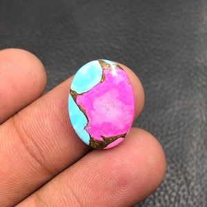 Può includere: Una gemma ovale con una tavolozza di colori vivaci. La pietra presenta una sorprendente combinazione di azzurro, rosa acceso e venature color oro. La superficie lucida riflette la luce, evidenziando gli esclusivi motivi cromatici.