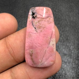Puede incluir: Una gema rectangular pulida con un color base rosa, con vetas rosas y negras más oscuras. La piedra tiene una superficie lisa y brillante y se sostiene en una mano. Esta gema probablemente se utiliza para joyería o fines decorativos.