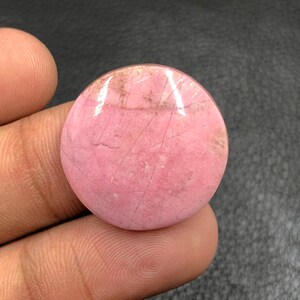 Puede incluir: Una gema redonda y pulida de color rosa, con sutiles inclusiones marrones y grises y finas líneas. La piedra se sostiene en una mano, mostrando su forma lisa y circular. Esta gema podría usarse para joyería.