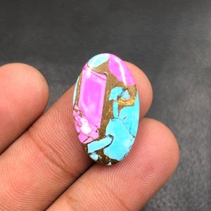 Può includere: Una gemma ovale con una tavolozza di colori vivaci. La pietra presenta una sorprendente combinazione di tonalità rosa, turchese e oro. Le sezioni rosa e turchese sono separate da venature dorate, creando un motivo unico. La gemma è tenuta in una mano.