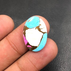 Può includere: Una gemma ovale con una tavolozza di colori vivaci. La pietra presenta un mix di motivi a matrice turchese, bianchi, rosa e dorati. La gemma è tenuta in una mano, mostrando la sua superficie lucida e il suo design unico.