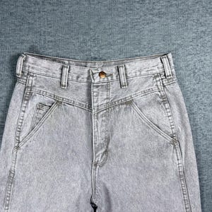 Peut inclure: Gros plan sur un jean en denim gris d&eacute;lav&eacute; &agrave; l'acide. Le jean a une taille haute, une fermeture boutonn&eacute;e et une coupe classique &agrave; cinq poches. Le tissu a un aspect textur&eacute; et les coutures sont visibles.