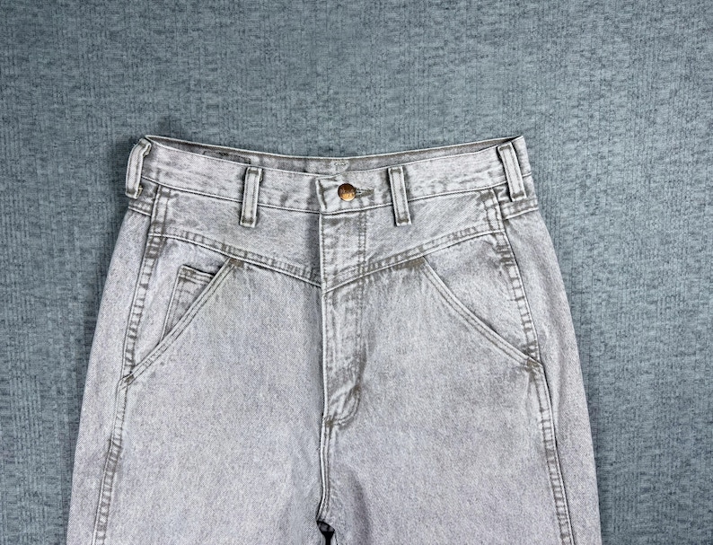 Peut inclure: Gros plan sur un jean en denim gris d&eacute;lav&eacute; &agrave; l'acide. Le jean a une taille haute, une fermeture boutonn&eacute;e et une coupe classique &agrave; cinq poches. Le tissu a un aspect textur&eacute; et les coutures sont visibles.