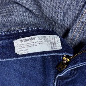 Pode incluir: Close-up de jeans Wrangler azuis. A etiqueta mostra "31 MWSDD 13/14 x 32" e "Made in USA 100% Cotton". Um z&iacute;per dourado &eacute; vis&iacute;vel.