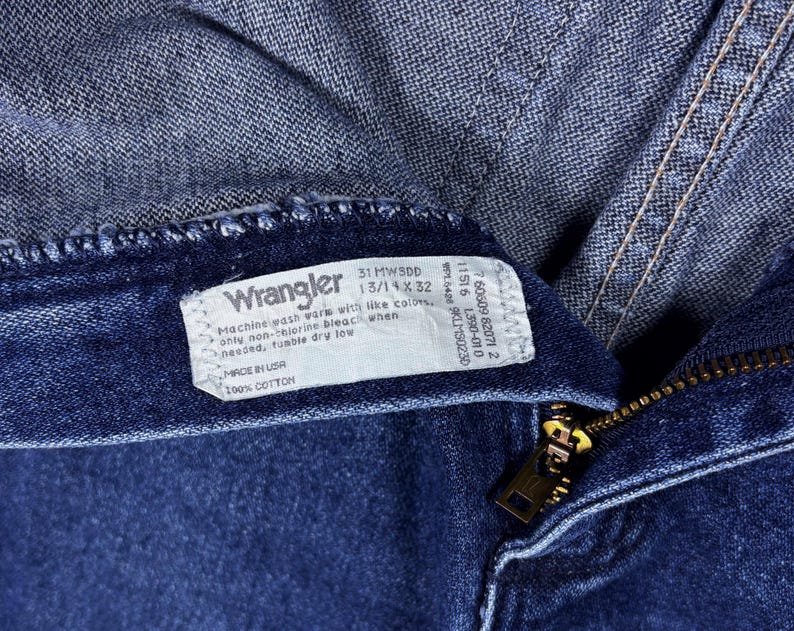 Pode incluir: Close-up de jeans Wrangler azuis. A etiqueta mostra "31 MWSDD 13/14 x 32" e "Made in USA 100% Cotton". Um z&iacute;per dourado &eacute; vis&iacute;vel.