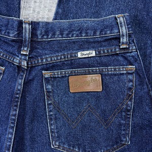 Pode incluir: Close-up de jeans de ganga azul escuro. O bolso traseiro apresenta um remendo de couro castanho com o nome da marca "Wrangler" em relevo. Os jeans t&ecirc;m um design cl&aacute;ssico de cinco bolsos com costuras vis&iacute;veis.