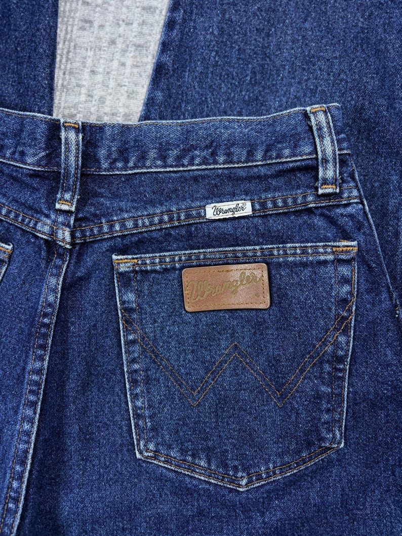 Pode incluir: Close-up de jeans de ganga azul escuro. O bolso traseiro apresenta um remendo de couro castanho com o nome da marca "Wrangler" em relevo. Os jeans t&ecirc;m um design cl&aacute;ssico de cinco bolsos com costuras vis&iacute;veis.