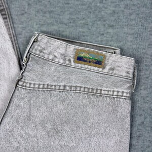 Peut inclure: Gros plan sur un jean en denim gris clair. Le jean a un aspect d&eacute;lav&eacute; et une &eacute;tiquette Wrangler sur la ceinture arri&egrave;re. L'&eacute;tiquette pr&eacute;sente une sc&egrave;ne de montagne avec le logo Wrangler. Le jean est pli&eacute;.