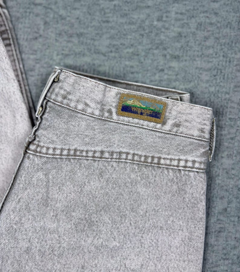 Peut inclure: Gros plan sur un jean en denim gris clair. Le jean a un aspect d&eacute;lav&eacute; et une &eacute;tiquette Wrangler sur la ceinture arri&egrave;re. L'&eacute;tiquette pr&eacute;sente une sc&egrave;ne de montagne avec le logo Wrangler. Le jean est pli&eacute;.