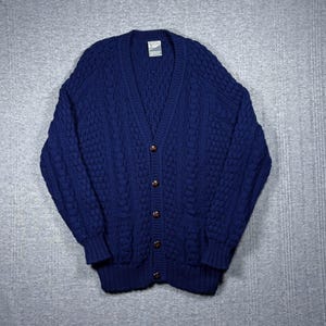 May include: A navy blue cable-knit cardigan with a V-neck and brown buttons. The sweater features a textured pattern and two front pockets. The garment is made of a thick, warm material.