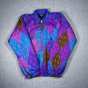 May include: A vibrant, patterned jacket featuring a striking geometric design in shades of purple, blue, and brown. The jacket has a collared neckline, a full zipper, and elastic cuffs and hem. The design is a mix of diamond and stripe patterns.
