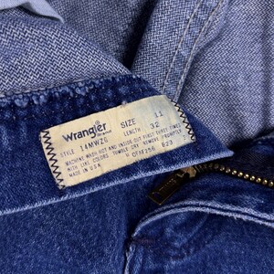 May include: Close-up of blue denim jeans with a Wrangler brand tag. The tag indicates size 11, length 32, and style 14MWZG. The jeans have a zipper and a button closure. The tag also includes washing instructions.