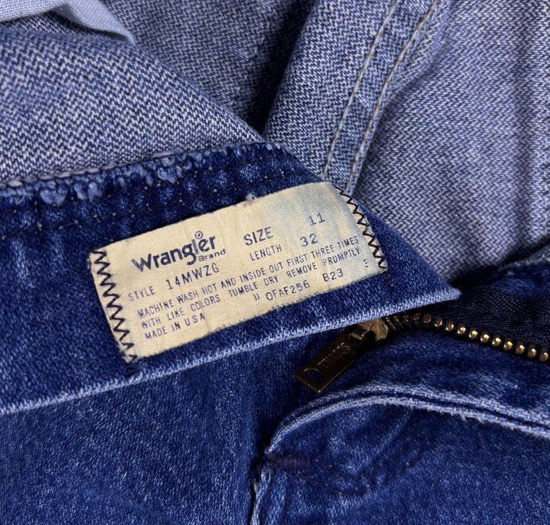 May include: Close-up of blue denim jeans with a Wrangler brand tag. The tag indicates size 11, length 32, and style 14MWZG. The jeans have a zipper and a button closure. The tag also includes washing instructions.