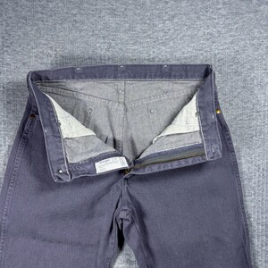 May include: Close-up of a pair of blue jeans with the zipper open. The jeans are a solid blue color with a gray interior. The jeans are laid flat on a gray surface.