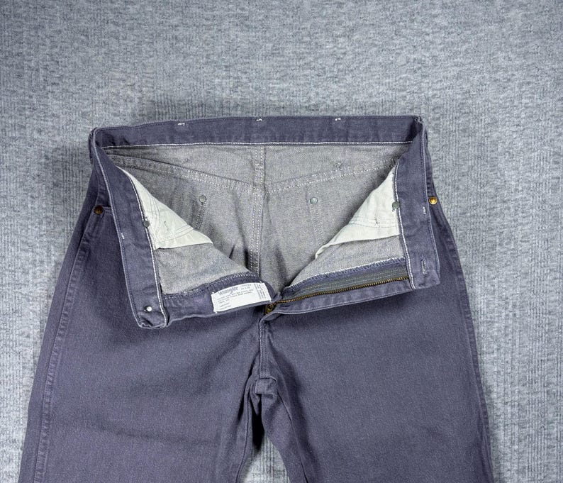 May include: Close-up of a pair of blue jeans with the zipper open. The jeans are a solid blue color with a gray interior. The jeans are laid flat on a gray surface.