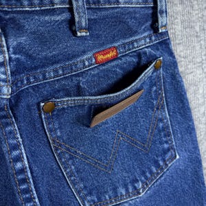 May include: Close-up of the back pocket of blue denim jeans. The pocket features a brown leather patch and a stitched 'W' design. The Wrangler label is visible on the waistband. The jeans have a classic, worn look.