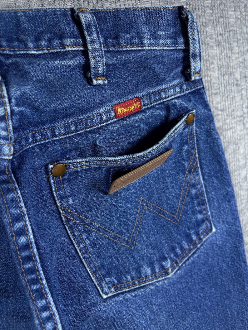 May include: Close-up of the back pocket of blue denim jeans. The pocket features a brown leather patch and a stitched 'W' design. The Wrangler label is visible on the waistband. The jeans have a classic, worn look.