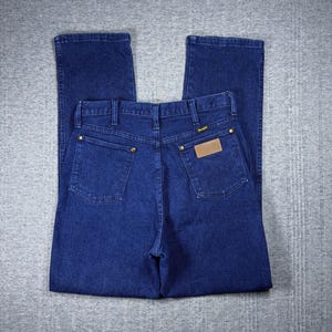 May include: Dark blue denim jeans with a classic five-pocket design. The jeans feature a straight leg cut, brass-colored rivets, and a brown leather patch with the brand name. The back pockets are square, and the jeans are folded neatly.