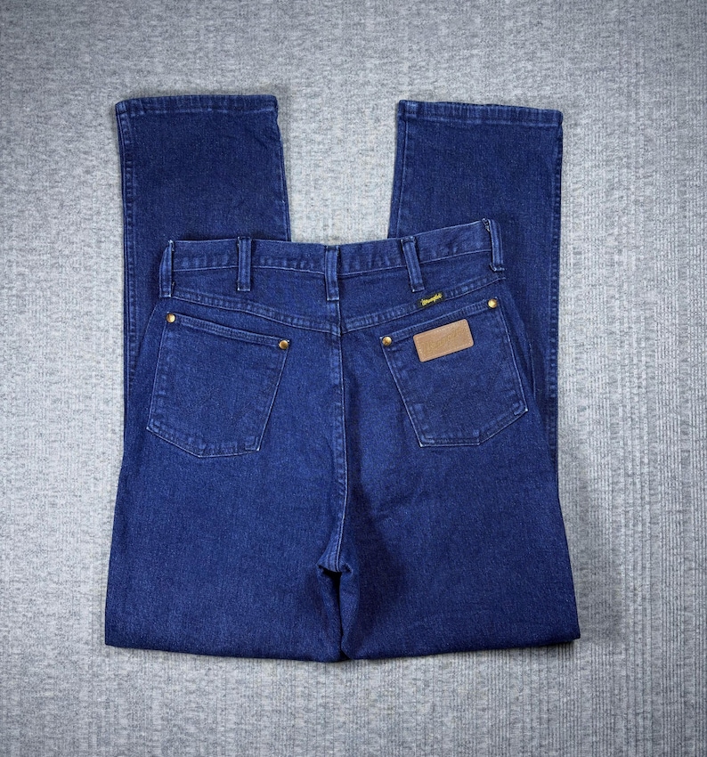 May include: Dark blue denim jeans with a classic five-pocket design. The jeans feature a straight leg cut, brass-colored rivets, and a brown leather patch with the brand name. The back pockets are square, and the jeans are folded neatly.