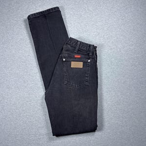 Könnte beinhalten: Schwarze Jeans aus Denim mit klassischem Fünf-Taschen-Design. Die Jeans haben einen braunen Lederaufnäher und ein rotes Logo-Etikett. Die Jeans liegen flach auf einer grauen Oberfläche.