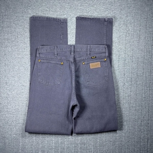 May include: A pair of blue-grey Wrangler jeans, viewed from the back. The jeans have two back pockets, gold rivets, and a brown leather patch with the Wrangler logo. The jeans are folded neatly on a grey surface.
