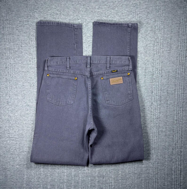 May include: A pair of blue-grey Wrangler jeans, viewed from the back. The jeans have two back pockets, gold rivets, and a brown leather patch with the Wrangler logo. The jeans are folded neatly on a grey surface.