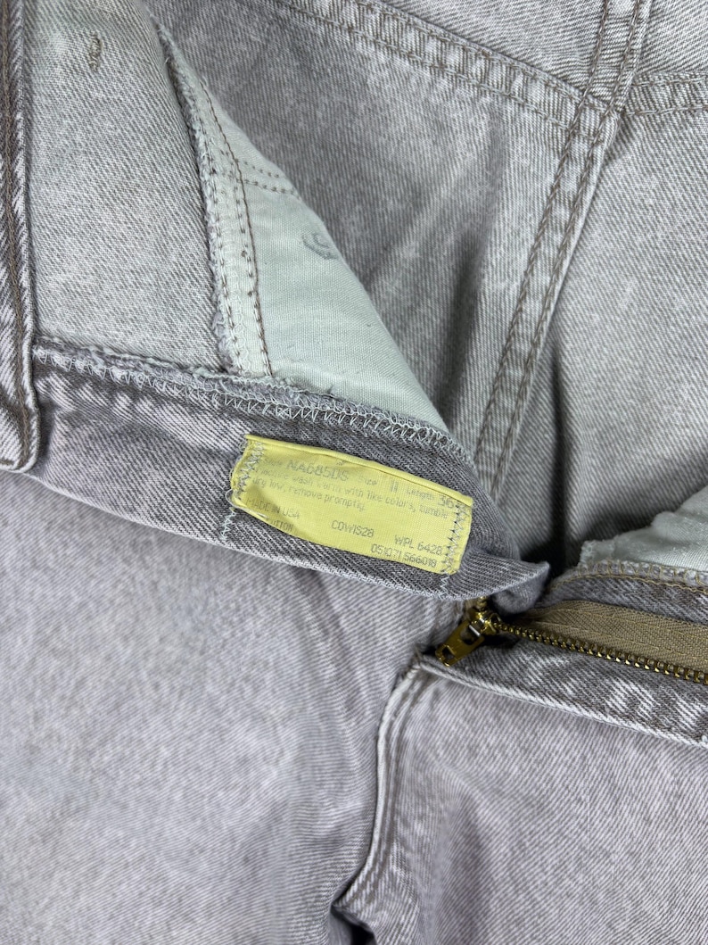 Peut inclure: Gros plan sur un jean en denim gris clair. Le jean a une fermeture &eacute;clair et une &eacute;tiquette jaune avec du texte. Le tissu semble us&eacute;, avec des coutures et des coutures visibles. Le style g&eacute;n&eacute;ral est d&eacute;contract&eacute;.