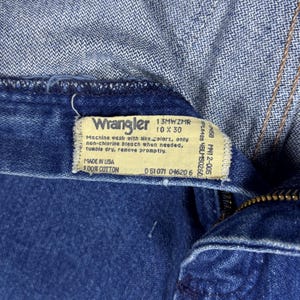 K&ouml;nnte beinhalten: Nahaufnahme einer blauen Wrangler-Jeans. Das Etikett zeigt "Wrangler 10 x 30" und "Made in USA 100% Cotton". Waschhinweise sind ebenfalls zu sehen. Die Jeans ist teilweise aufgekn&ouml;pft und zeigt den Rei&szlig;verschluss.