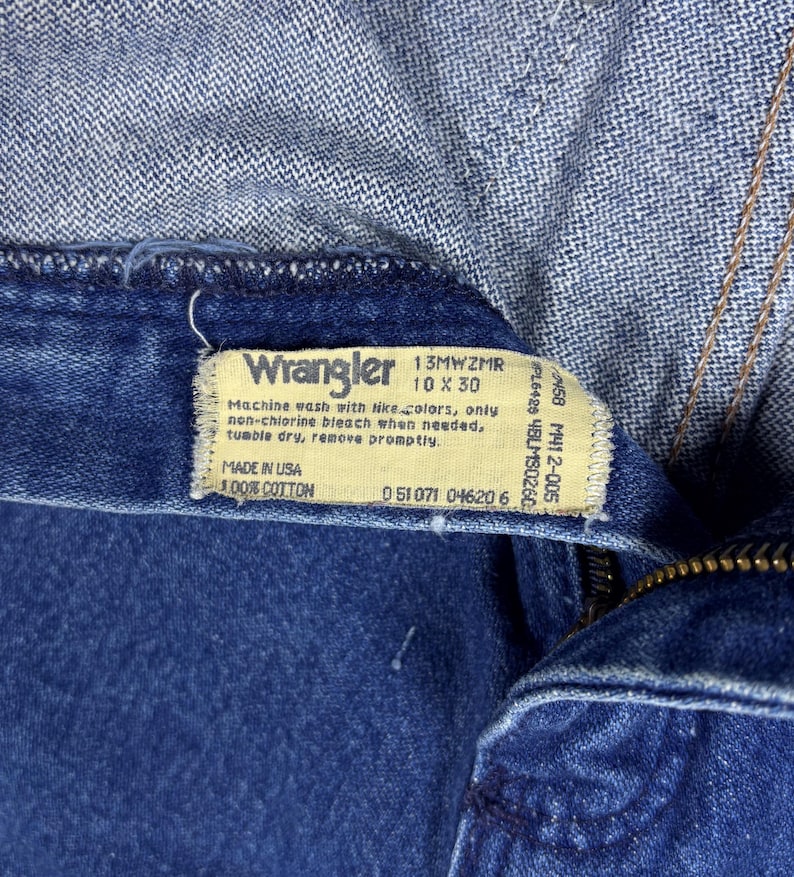 K&ouml;nnte beinhalten: Nahaufnahme einer blauen Wrangler-Jeans. Das Etikett zeigt "Wrangler 10 x 30" und "Made in USA 100% Cotton". Waschhinweise sind ebenfalls zu sehen. Die Jeans ist teilweise aufgekn&ouml;pft und zeigt den Rei&szlig;verschluss.