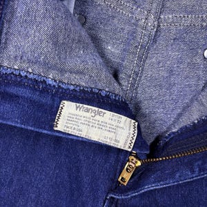 May include: Close-up of a pair of dark blue Wrangler jeans. The jeans have a zipper and a label with washing instructions and size information. The label reads "Wrangler 13MTIM 14X32".