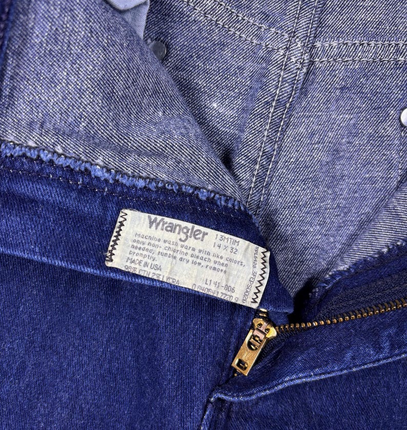 May include: Close-up of a pair of dark blue Wrangler jeans. The jeans have a zipper and a label with washing instructions and size information. The label reads "Wrangler 13MTIM 14X32".