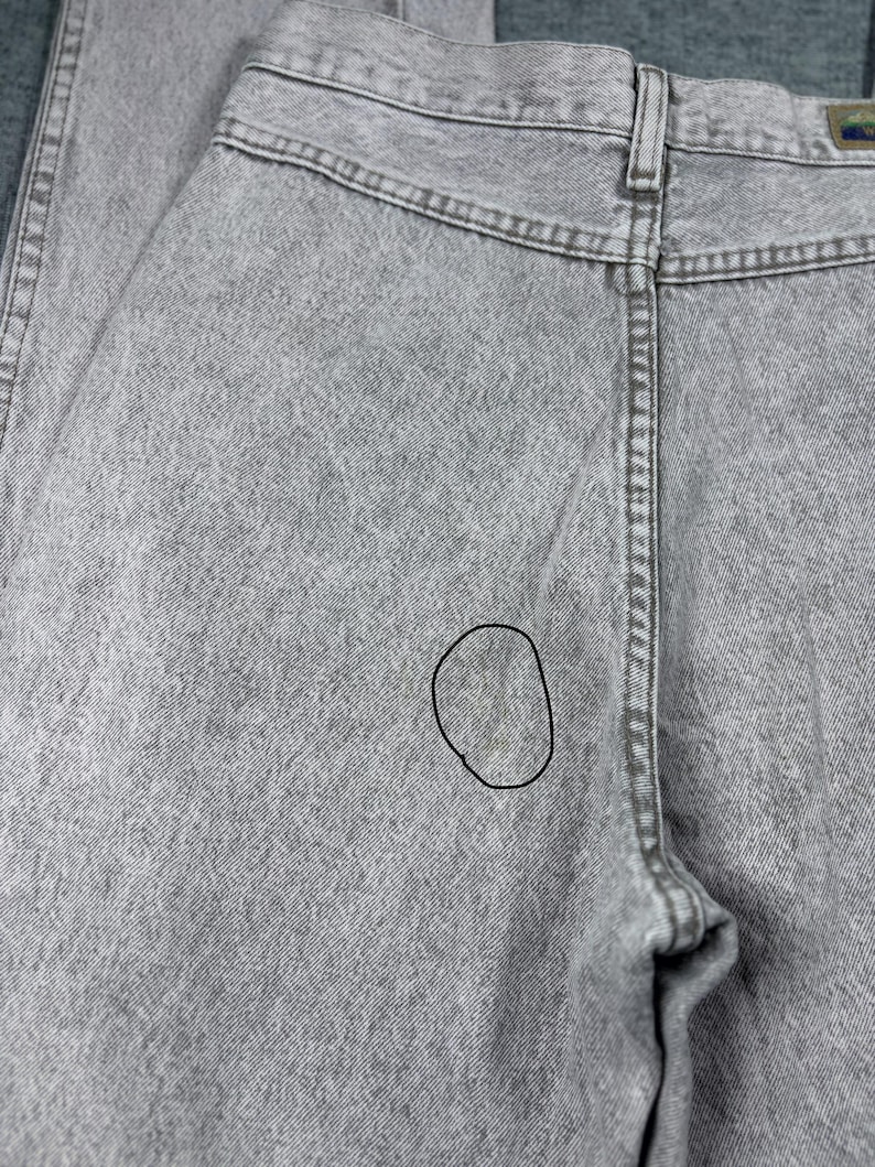 Peut inclure: Gros plan du dos d'un jean en denim gris clair. Le jean a une conception classique &agrave; cinq poches avec une fermeture &agrave; bouton et &agrave; glissi&egrave;re. Le tissu a un aspect textur&eacute; et il y a une petite tache au dos.