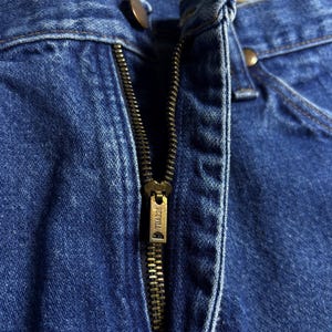 May include: Close-up of blue denim jeans with a partially open brass zipper. The zipper pull is marked with the brand name "SCOVILL". Two brass buttons are visible near the waistband.