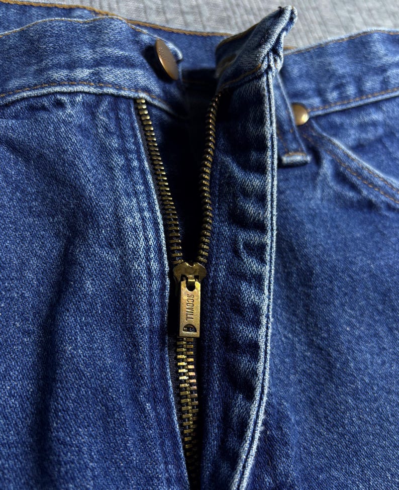 May include: Close-up of blue denim jeans with a partially open brass zipper. The zipper pull is marked with the brand name "SCOVILL". Two brass buttons are visible near the waistband.