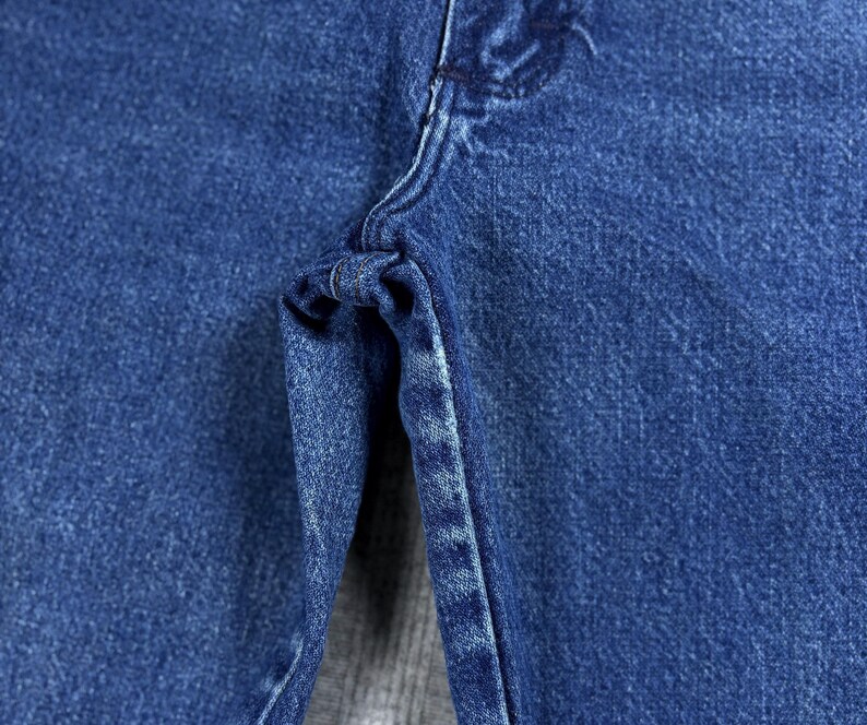May include: Close-up of blue denim jeans, showcasing the texture and stitching details. The image highlights the fabric's weave and the construction of the garment, focusing on the seam and the area around the zipper.