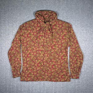 Könnte beinhalten: Eine langärmlige Bluse mit hohem Kragen und Bindedetail. Die Bluse hat ein Muster aus roten Beeren und grünen Blättern auf beigem Hintergrund. Der Stoff scheint leicht zu sein.