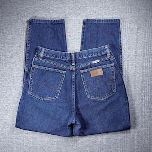 Pode incluir: Jeans de ganga azul escuro com um design cl&aacute;ssico de cinco bolsos. Os jeans apresentam um remendo de couro castanho com o nome da marca e um logotipo costurado nos bolsos traseiros. Os jeans est&atilde;o colocados sobre uma superf&iacute;cie cinzenta.