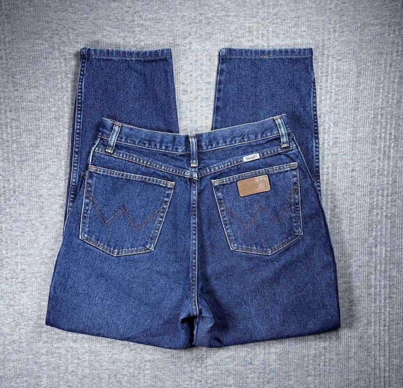 Pode incluir: Jeans de ganga azul escuro com um design cl&aacute;ssico de cinco bolsos. Os jeans apresentam um remendo de couro castanho com o nome da marca e um logotipo costurado nos bolsos traseiros. Os jeans est&atilde;o colocados sobre uma superf&iacute;cie cinzenta.