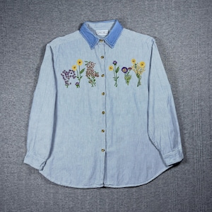 Op de afbeelding: Een overhemd met lange mouwen en een lichtblauw en wit gestreept patroon. Het heeft een denim kraag en geborduurde bloemen in verschillende kleuren over de borst. De kraag toont de merknaam "Oak Hill".