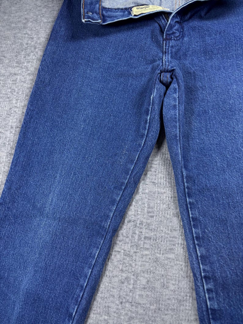 K&ouml;nnte beinhalten: Nahaufnahme einer blauen Jeans. Die Jeans hat ein klassisches F&uuml;nf-Taschen-Design, einen Knopf- und Rei&szlig;verschluss und der Markenname "Wrangler" ist auf dem Bund sichtbar. Der Denim hat ein leicht verwaschenes Aussehen.