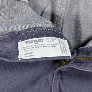 May include: Close-up of a pair of gray Wrangler jeans. The label reads "Wrangler 34 x 38" and includes washing instructions. The jeans are made of 100% cotton and have a brass zipper.