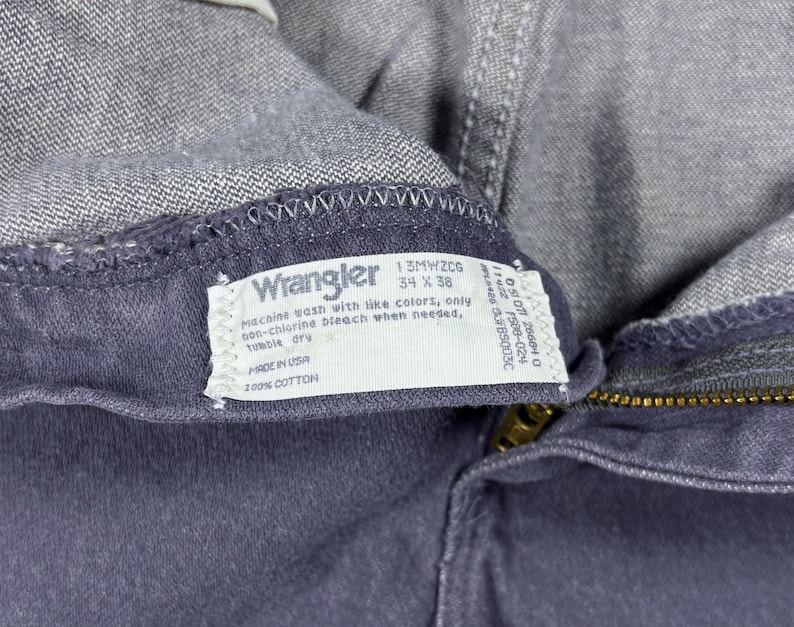 May include: Close-up of a pair of gray Wrangler jeans. The label reads "Wrangler 34 x 38" and includes washing instructions. The jeans are made of 100% cotton and have a brass zipper.