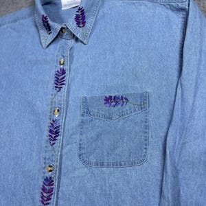 May include: Light blue denim shirt with long sleeves. The shirt features embroidered purple lavender sprigs along the collar, button placket, and pocket. Gold-colored buttons secure the front. A classic, casual top.