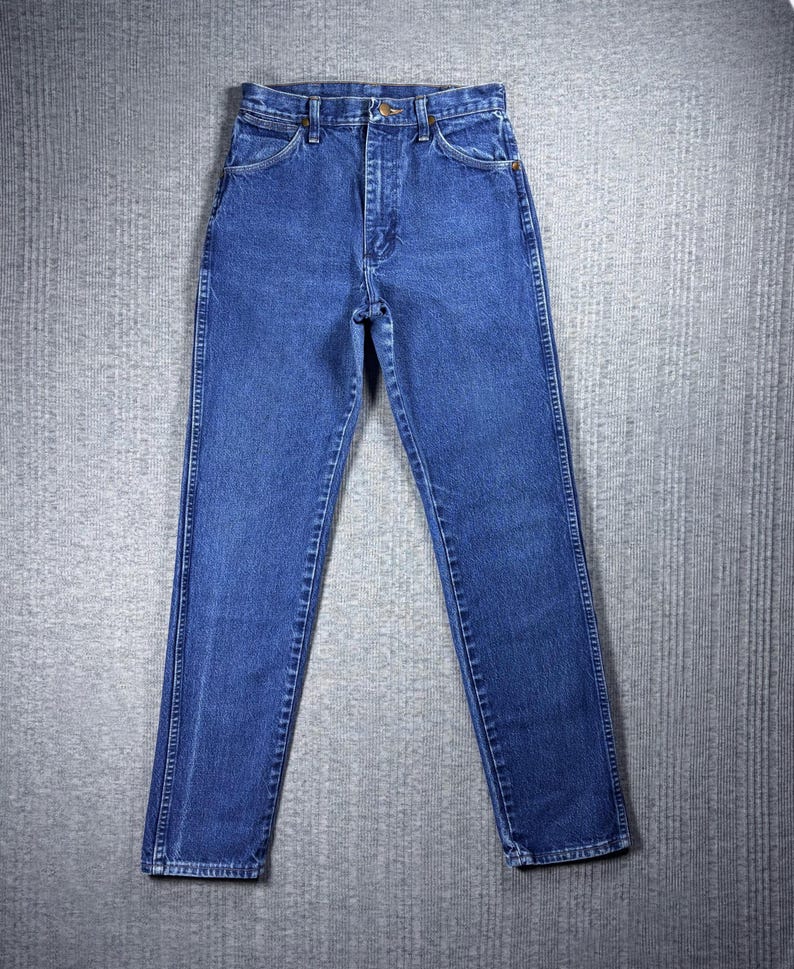 May include: Classic blue denim jeans with a straight leg cut. The jeans feature a button and zipper closure, front and back pockets, and belt loops. The denim is a medium wash, and the jeans are laid flat on a gray surface.