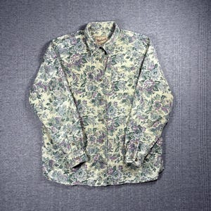 May include: A button-down shirt with a floral pattern in shades of green, purple, and cream. The long-sleeved shirt has a collared neckline and a single chest pocket. The Woolrich brand logo is visible on the collar.